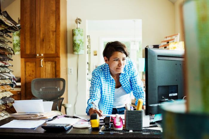 Mulher trabalhando em home office, analisando documentos e olhando para o computador. A imagem representa os desafios e soluções sobre como gerenciar equipes home office, com foco em produtividade e organização.