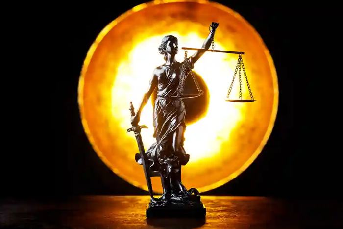 Estátua da Justiça em metal escuro iluminada por um círculo dourado intenso ao fundo, segurando balança e espada, em composição que sugere a travessia além das formas empresariais para alcançar responsabilidades reais, alinhada ao conceito de desconsideração da personalidade jurídica em licitações, onde o direito ultrapassa aparências societárias e incide sobre o núcleo efetivo do poder e da decisão.